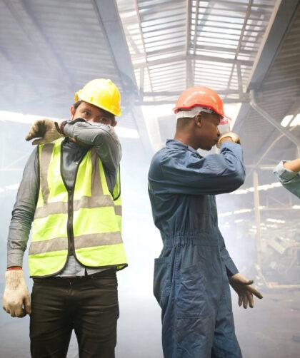 Fabrikarbeiter stehen in Rauch und Abgasen, halten sich die Nase zu.